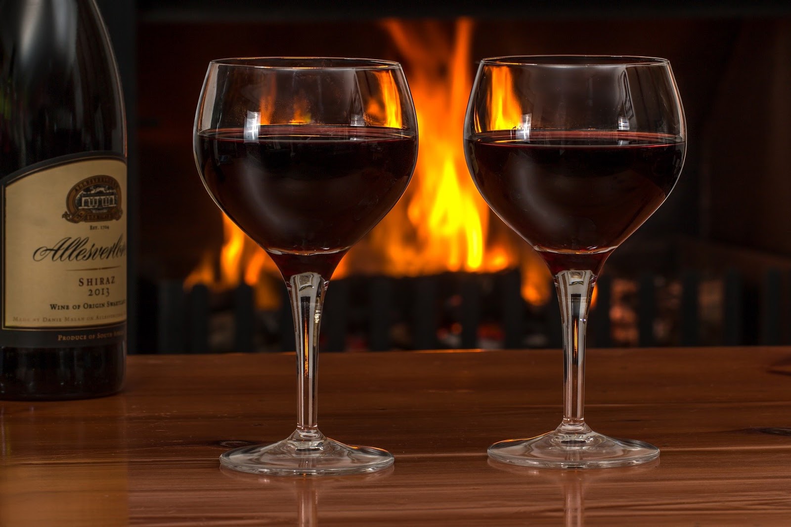 A bottle and two glasses of wine by a fire.